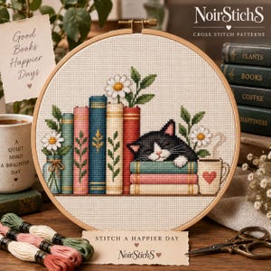 May include: A circular wooden embroidery hoop containing a cross-stitch pattern of a black and white cat sleeping on a stack of colourful books. The design includes daisies, a mug with a heart, and the text Good Books Happier Days on a paper note.