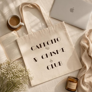 May include: Cream-colored tote bag with the words "Cafecito & Chisme Club" in brown lettering. The bag is on a beige surface, alongside a cup of coffee, a book, a laptop, a candle, and flowers.