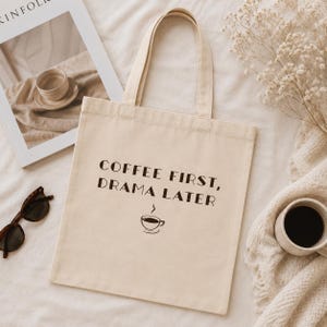 May include: Cream-colored tote bag with the text "COFFEE FIRST, DRAMA LATER" in black. A small coffee cup graphic is below the text. The bag has two handles and is sitting on a white surface.