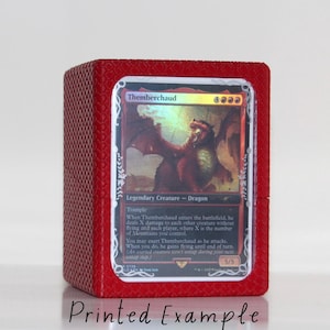 Puede incluir: Una caja roja texturizada, impresa en 3D, con una carta de Magic: The Gathering en su interior. La carta presenta un dragón rojo con un efecto holográfico y el texto "Themberchaud". La carta es una criatura legendaria - dragón con una fuerza de 5 y una resistencia de 5.