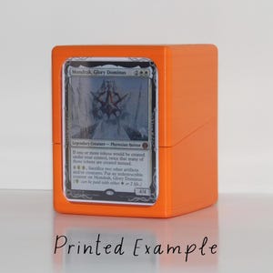 Op de afbeelding: Oranje plastic deckbox met een zwart-witte Magic: The Gathering kaart waarop Mondrak, Glory Dominus te zien is. De tekst op de kaart luidt: "Als een of meer tokens onder jouw controle zouden worden gecreëerd, worden er twee keer zoveel van die tokens in plaats daarvan gecreëerd. 4, Offer twee andere artefacten en/of wezens: Plaats een onverwoestbare teller op Mondrak, Glory Dominus. ( kan worden betaald met of 2 levens.) 4/4".