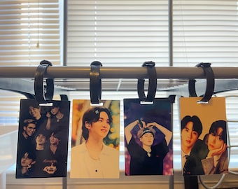 May include: Four luggage tags with different images of a K-pop band. The tags are hanging from a metal rack. The images are of the band members in various poses and outfits. The tags are made of a white plastic material.