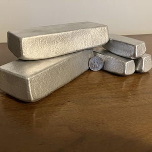 Puede incluir: Una colección de barras metálicas rectangulares de color plateado, algunas apiladas, con una moneda como escala. Las barras tienen una superficie texturizada y están dispuestas sobre una superficie de madera. Las barras parecen ser de metal fundido.