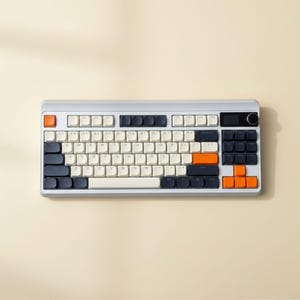 May include: A mechanical keyboard with a light gray frame, featuring a mix of cream, dark gray, and bright orange keycaps. The layout includes a standard QWERTY arrangement, a dedicated number pad, a small digital display, and a circular knob.