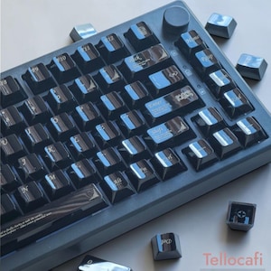 Op de afbeelding: Een donkergrijs mechanisch toetsenbord met glanzende zwarte toetsen, witte letters en decoratieve filigraanaccenten. Het toetsenbord heeft een ronde volumeknop in de rechterbovenhoek en er liggen enkele losse toetsen rondom de basis.