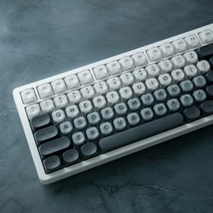 May include: A mechanical keyboard featuring a white frame and a gradient color scheme on the keycaps, transitioning from white on the left and top to dark gray on the right and bottom. The keys display standard QWERTY layout with black lettering.
