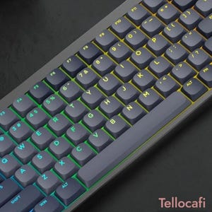 Puede incluir: Un teclado mecánico gris oscuro con distribución QWERTY y teclas retroiluminadas. Las letras y símbolos de las teclas están iluminados por un degradado de luz que transiciona del azul en el lado izquierdo al amarillo en el lado derecho.