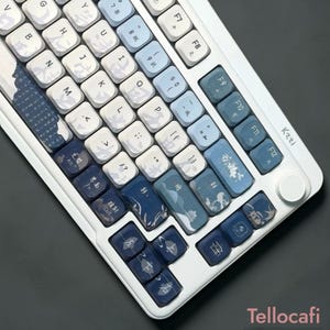以下が含まれることがあります： 白いメカニカルキーボード、カスタムキーキャップ付き。キーキャップは、青と白のグラデーションで、日本風のアートワークが特徴です。キーボードは白いフレームとKzziのロゴが付いています。
