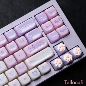 May include: A custom mechanical keyboard with a lavender and white color scheme. The keycaps feature a variety of designs, including paw prints, a cat illustration, and the text "Backspace." The keyboard has a modern, minimalist aesthetic.