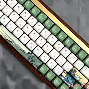 Könnte beinhalten: Eine mechanische Tastatur mit einem dunklen Holzrahmen, ausgestattet mit weißen Tasten mit roten lateinischen Buchstaben und japanischen Schriftzeichen sowie salbeigrünen Tasten. Die Leertaste und die Eingabetaste zeigen eine Landschaftsillustration.
