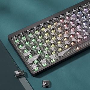 Puede incluir: Un teclado mecánico negro con teclas transparentes, letras blancas e iluminación RGB integrada que brilla en tonos verdes, amarillos y morados. Dos teclas negras sueltas con las etiquetas Ctrl y Del están situadas cerca del teclado.