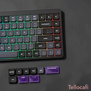 May include: A black mechanical keyboard with backlit keys featuring a gradient of green, yellow, and red light. Below the keyboard are several loose keycaps, including black ones labeled option, fn, and del, and translucent purple keycaps labeled backspace, tab, and a slash symbol.