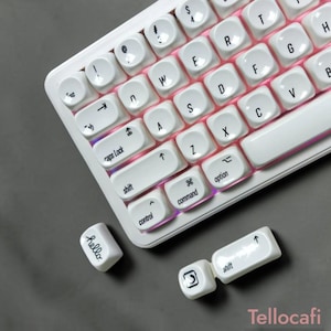 Puede incluir: Un teclado mecánico blanco con teclas redondeadas, letras negras e iluminación rosa. Dos teclas blancas sueltas están colocadas cerca, una con la palabra hello y la otra con un icono de flecha y la palabra shift.