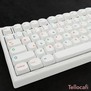 Może przedstawiać: Biała klawiatura mechaniczna z białymi nakładkami na klawisze. Nakładki na klawisze mają kolorowe litery i symbole. Klawiatura ma minimalistyczny design i jest ustawiona na czarnym tle. Słowo "Tellocafi" jest widoczne w prawym dolnym rogu.