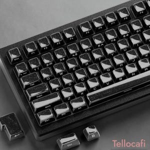 May include: A black mechanical keyboard featuring keycaps with a subtle glittery finish. The keys display standard QWERTY layout characters in white. Three loose keycaps with the same dark, sparkling texture are positioned near the keyboard base.