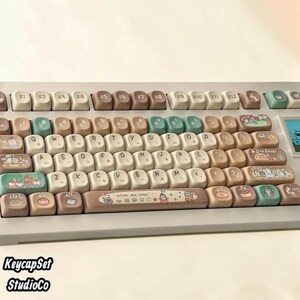 Könnte beinhalten: Eine mechanische Tastatur mit einem benutzerdefinierten Tastenkappenset in einer Palette aus Creme, Braun und gedämpftem Blaugrün. Die Tasten zeigen verschiedene Illustrationen von Lebensmitteln und kleinen Figuren, mit dem Text autumn day comes.