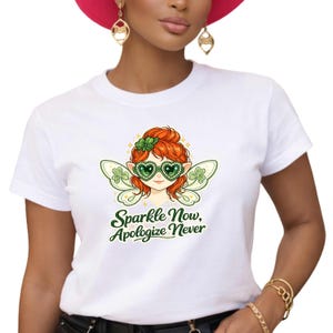 May include: White t-shirt with a cartoon fairy design. The fairy has red hair, green heart-shaped glasses, and shamrock wings. The text "Sparkle Now, Apologize Never" is printed in green script. The t-shirt has a crew neck.