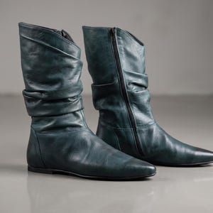 May include: A pair of dark teal mid-calf boots crafted from smooth leather. These boots feature a slouchy, gathered shaft design, a pointed toe, a flat sole, and a black side zip closure that runs along the inner side of each boot.