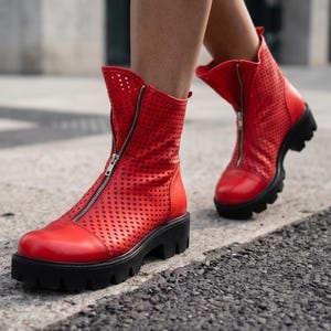 May include: A pair of bright red ankle boots featuring a perforated upper design, a front-facing silver zipper closure, and a thick, black lugged rubber sole. The boots have a rounded toe and a structured shaft that extends above the ankle.