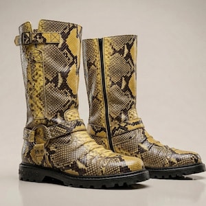 Könnte beinhalten: Ein Paar hohe Stiefel mit gelbem und braunem Schlangenmuster. Die Stiefel haben eine schwarze Sohle, einen Schnallenverschluss und einen Reißverschluss an der Seite. Die Stiefel sind kniehoch.