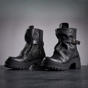 May include: A pair of black ankle boots featuring a slouchy, gathered shaft, a side zip closure, and a horizontal strap with a metallic buckle across the mid-section. The boots have a thick, lugged rubber sole and a block heel.