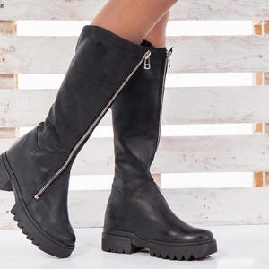 Könnte beinhalten: Ein Paar kniehohe schwarze Stiefel mit einem auffälligen, silberfarbenen Frontreißverschluss, der vertikal am Schaft verläuft. Die Stiefel haben eine abgerundete Zehenpartie, eine dicke Profilsohle mit tiefem Muster und einen flachen Blockabsatz.