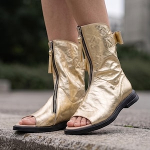 May include: Gold metallic open-toe boots with black soles and zippers. The boots have a mid-calf height and a small bow detail on the side. The boots are made of a shiny, textured material.