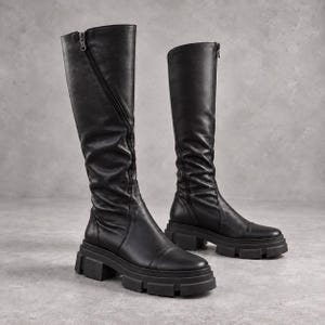 Puede incluir: Un par de botas negras hasta la rodilla con acabado liso, punta redondeada y suela gruesa y dentada. Las botas cuentan con un cierre de cremallera lateral vertical y detalles fruncidos a lo largo de la caña para una apariencia texturizada.