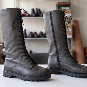 Könnte beinhalten: Ein Paar kniehohe graue Lederstiefel mit schwarzer Schnürung an der Vorderseite, seitlichem Reißverschluss, abgerundeter Zehenkappe und einer dicken, profilierten schwarzen Gummisohle. Die Stiefel haben eine glatte Oberfläche und einen hohen Schaft.