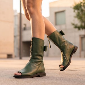 May include: Green leather open-toe boots with a zipper closure and a small bow detail. The boots have a flat sole and are mid-calf height. The boots are a solid green color.