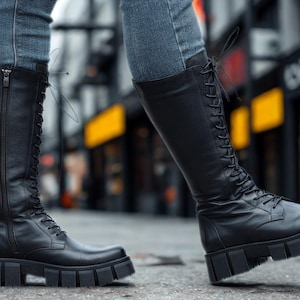 May include: Black knee-high boots with a lace-up design and chunky soles. The boots have a zipper closure on the side and are worn with blue jeans. The boots are made of a smooth, matte material.