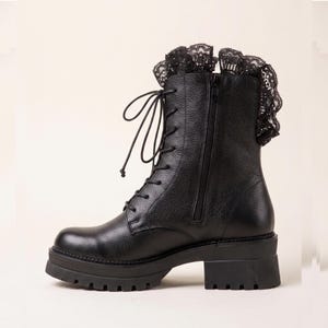 Könnte beinhalten: Ein schwarzer Stiefeletten-Stiefel aus strukturiertem Leder mit Schnürung vorne, passenden schwarzen Schnürsenkeln, seitlichem Reißverschluss, dicker Profilsohle mit Blockabsatz und einem dekorativen schwarzen Spitzenrüschenbesatz am Schaft.