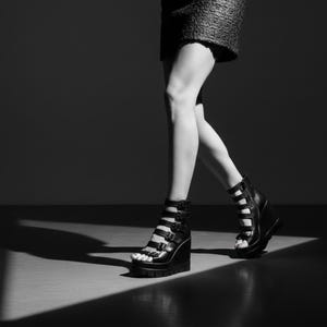 May include: Black and white image of platform sandals with multiple straps and buckles. The sandals have a wedge heel and are worn by a person walking. The image is in a studio setting with dramatic lighting.