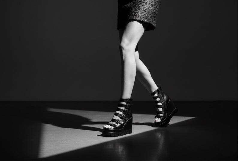 May include: Black and white image of platform sandals with multiple straps and buckles. The sandals have a wedge heel and are worn by a person walking. The image is in a studio setting with dramatic lighting.