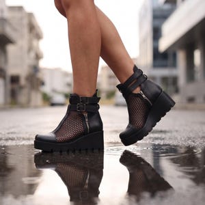 Könnte beinhalten: Schwarze Leder-Wedge-Ankle-Boots mit Mesh-Einsätzen und Reißverschluss. Die Stiefel haben eine Plateausohle und verstellbare Riemen mit Schnallen. Das Schuhwerk wird im Freien auf einer nassen Oberfläche gezeigt.