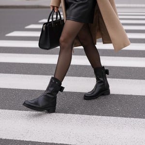 Könnte beinhalten: Eine Person überquert einen Zebrastreifen und trägt schwarze Lederstiefel mit seitlichen Schnallen, einen schwarzen Rock und einen beigefarbenen Mantel. Die Person trägt eine schwarze Handtasche.