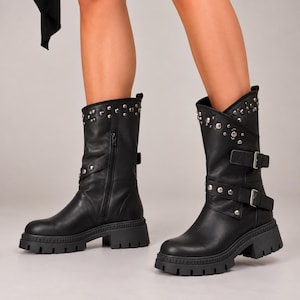 May include: Black mid-calf boots featuring a thick lug sole, a side zipper, and two adjustable straps with silver-toned metal buckles. The upper shaft is decorated with a row of silver-toned metal studs along the top edge and the straps.