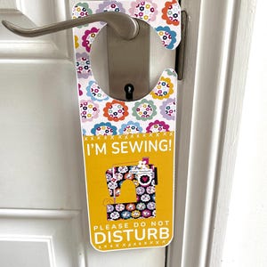 May include: A yellow door hanger with the text "I'M SEWING!" and "PLEASE DO NOT DISTURB." The hanger features a floral pattern and a sewing machine graphic. It is hanging on a white door with a silver handle.
