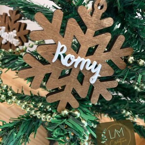 Peut inclure: Ornement de Noël en forme de flocon de neige en bois avec le nom "Romy" en écriture blanche. L'ornement est marron et est suspendu à une ficelle de jute. L'ornement est sur un sapin de Noël vert avec une guirlande de perles dorées.