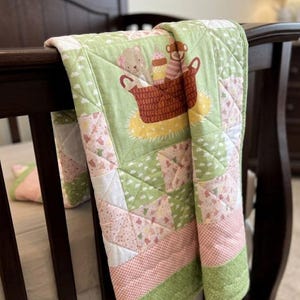 May include: A baby quilt draped over a dark brown crib. The quilt features a central panel with a brown basket containing a teddy bear and a monkey. The quilt has a green background with pink and white geometric patterns and a pink and white gingham border.