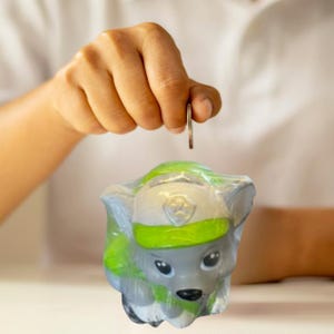 May include: A gray and green puppy-shaped piggy bank with a coin being dropped into the slot. The piggy bank has a white hat with a green stripe and a badge. The coin is held by a person's hand.