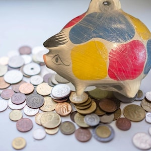 May include: A colorful ceramic piggy bank with patches of yellow, red, and blue. The piggy bank is surrounded by a collection of coins of various sizes and denominations, suggesting savings or financial planning.