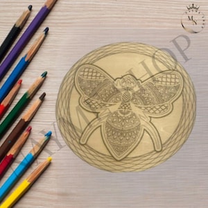 May include: A round, light brown wooden coaster with an engraved bee design. The bee is centered within a decorative border. Colored pencils in various colors are arranged along the left side of the image.