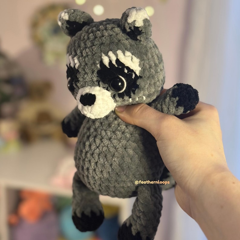 May include: A handmade gray and white crochet raccoon plush toy with black accents. The raccoon has large black eyes, a white muzzle, and black paws. The plush is soft and cuddly, perfect for a child's toy or nursery decor.