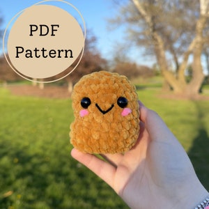 May include: A crocheted, golden-brown nugget toy with black eyes, a smiling mouth, and pink cheeks. The text "PDF Pattern" is displayed in a circular design. The nugget is held in a hand against a blurred outdoor background.