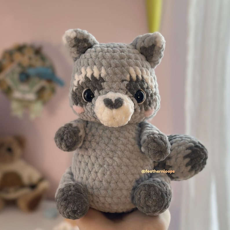 May include: A handmade crochet raccoon plushie in shades of gray, white, and black. The raccoon has a heart-shaped nose, pink cheeks, and a fluffy tail. The plushie is soft and cuddly, perfect for a child's toy or nursery decor.
