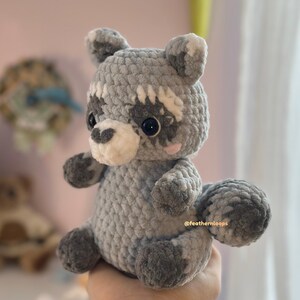 May include: A handmade crochet raccoon plushie in shades of gray and white. The stuffed animal has a round body, a fluffy tail, and a distinctive bandit mask around its eyes. The plushie is soft and cuddly, perfect for a child's toy or nursery decor.