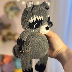 May include: A handmade crochet raccoon plush toy. The plush is gray with black and white accents, including a mask around the eyes and a striped tail. The text "@feathernloops" is visible on the plush.