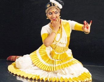 May include: A woman in a traditional Indian dance costume, featuring a white and yellow pleated skirt and top. The dancer wears gold jewelry, including a headpiece, necklace, and armbands. She is smiling and posed in a dance position.