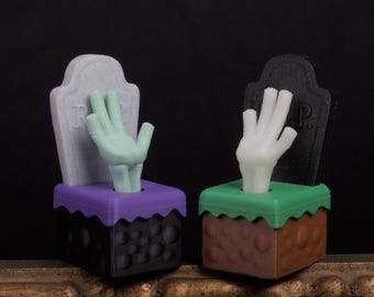 May include: Two Halloween-themed decorations featuring a hand emerging from a grave. One has a gray tombstone and a green hand, the other has a black tombstone and a white hand. The bases are brown and black.